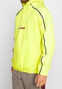 Neon gul pulloverjakke med lynlåse hætte, hvid og rød logo på forsiden og kontrasterende blå sidelåse. Har elastiske manchetter.