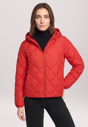 Femme aux cheveux bruns portant une veste matelassée rouge à capuche par-dessus un col roulé noir et un pantalon noir, debout avec une main sur la hanche.
