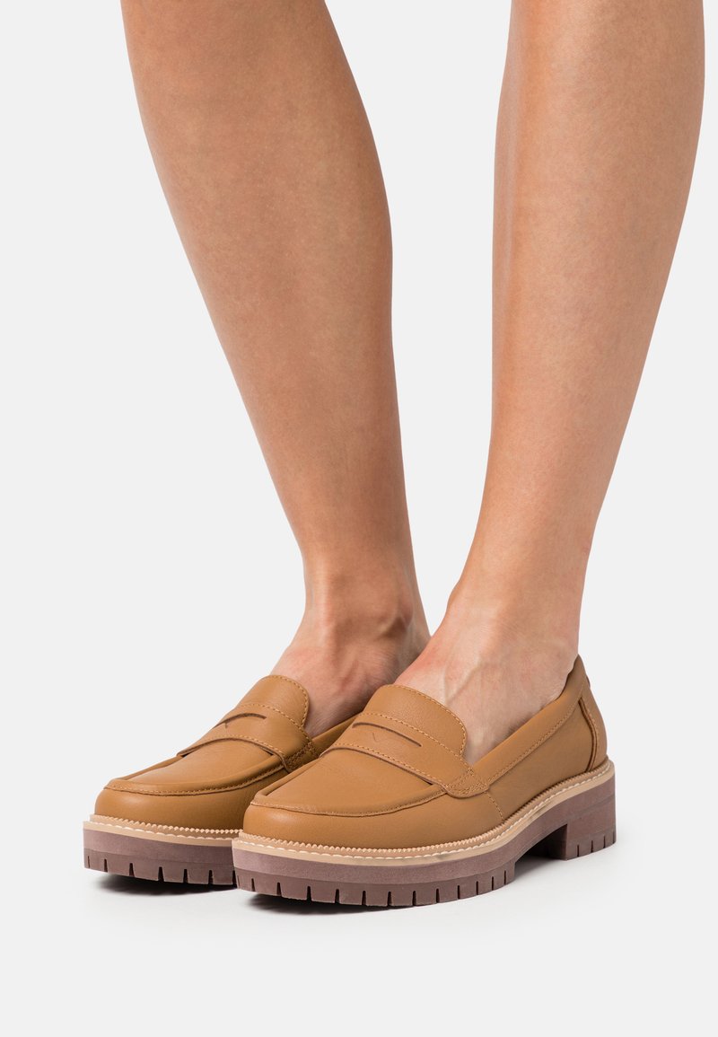 Mocasines de cuero color tan con punta redondeada, que presentan detalles de costura y una suela gruesa y estriada para mayor agarre y comodidad.