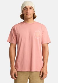 Pink cotton t-shirt with short sleeves and a crew neck, featuring a circular white logo on the left chest. Worn with beige corduroy pants.