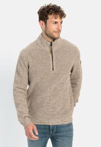 Beige trui met een hoge kraag en een halve rits aan de voorkant, met een gestructureerd gebreid patroon, geribbelde manchetten en een klein logo-opdruk op de linkermouw.