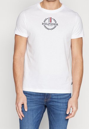 Weißes Baumwoll-T-Shirt mit einem kreisförmigen schwarzen und roten Logo mit Lorbeerakzenten. Kurze Ärmel und ein rundes Halsbündchen, kombiniert mit blauen Jeans.