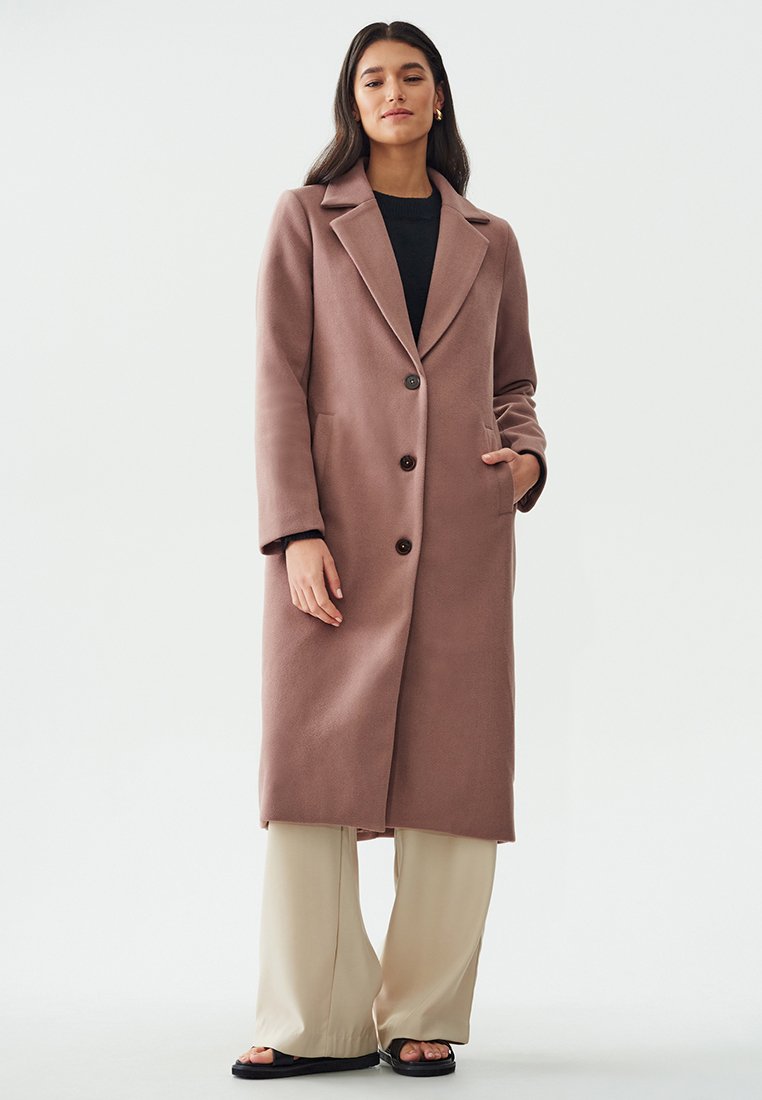 Lungo cappotto rosa realizzato in morbido tessuto, dotato di colletto a revers, due bottoni e tasche laterali. Abbinato a pantaloni beige a gamba larga.