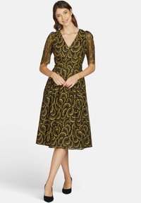 Schwarzes knielanges Kleid mit gelbem Wirbelmuster, V-Ausschnitt, durchsichtige Spitzenärmel, taillierte Passform und ausgestellter Rock. Schwarze Absatzschuhe.