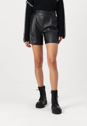 Modell trägt schwarze, hohe Fake-Leder-Shorts mit Knopfverschluss am Bund, kombiniert mit schwarzen, klobigen Schnürstiefeln und einem schwarzen Strickpullover.