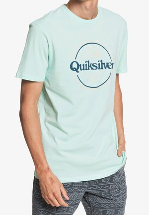 Mand iført lyseblå Quiksilver t-shirt og mønstrede mørke shorts, stående med den ene hånd i lommen og den anden afslappet ved siden.