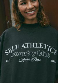 Felpa nera con audace testo bianco: "SELF ATHLETICS Country Club est. 2023 Sports Dept." Testo in vari stili di font. Tessuto testurizzato.