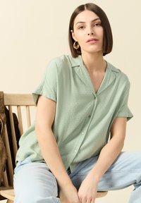 Lichtgroen shirt met korte mouwen, voorzien van een subtiel gestructureerd patroon, met knoopsluiting aan de voorkant en een kraag. Gecombineerd met lichtblauwe spijkerbroek.