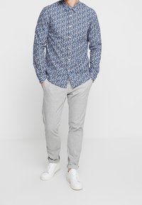 Homme portant une chemise boutonnée à fleurs bleues, un pantalon gris clair et des baskets blanches, se tenant les mains dans les poches.