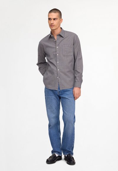 Homme portant une chemise à manches longues à carreaux marron et bleu, un jean bleu, des chaussettes blanches et des mocassins noirs, debout devant un fond blanc.