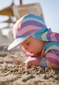 Värikäs raidallinen uimapaita ja myssy vaaleissa purppuran, sinisen ja vaaleanpunaisen sävyissä. Lapsi leikkii hiekkaisella pinnalla, keskittyneenä tekstuureihin.
