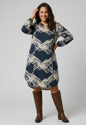 Femme souriante portant une robe bleu marine et beige à motifs, longueur aux genoux, manches longues, et des bottes marron montant aux genoux, debout, main sur la hanche.