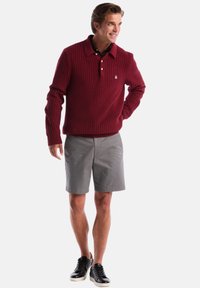 Bordeauxfarbener Strickpullover mit einem Kragen, kombiniert mit grauen Shorts und schwarzen Turnschuhen. Bemerkenswertes textiles Muster auf dem Pullover.
