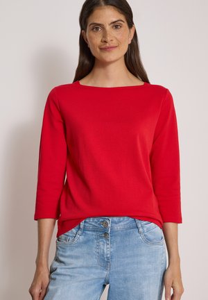 Femme aux longs cheveux foncés portant un haut rouge à manches longues et un jean bleu clair, debout devant un fond clair uni.