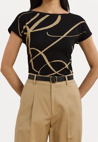 Svart kortärmad topp med gyllene abstrakta linjer, rund halsringning, tillsammans med beige högmidjade byxor och ett svart bälte med gyllene spänne.