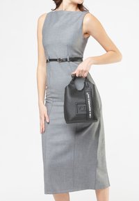 Donna in un vestito grigio aderente senza maniche con una cintura nera, che tiene una borsa nera con l'etichetta "IO SONO UNA BORSA RESPONSABILE."
