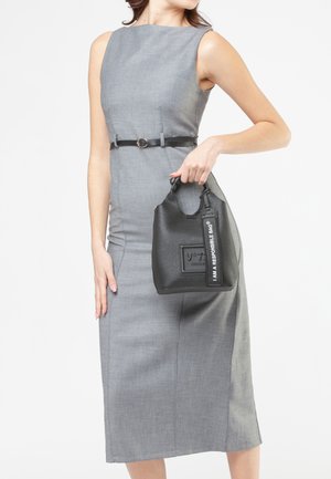 Vrouw in een grijze mouwloze jurk die goed aansluit met een zwarte riem, en een zwarte handtas vasthoudend met het label "IK BEN EEN VERANTWOORDE TAS."
