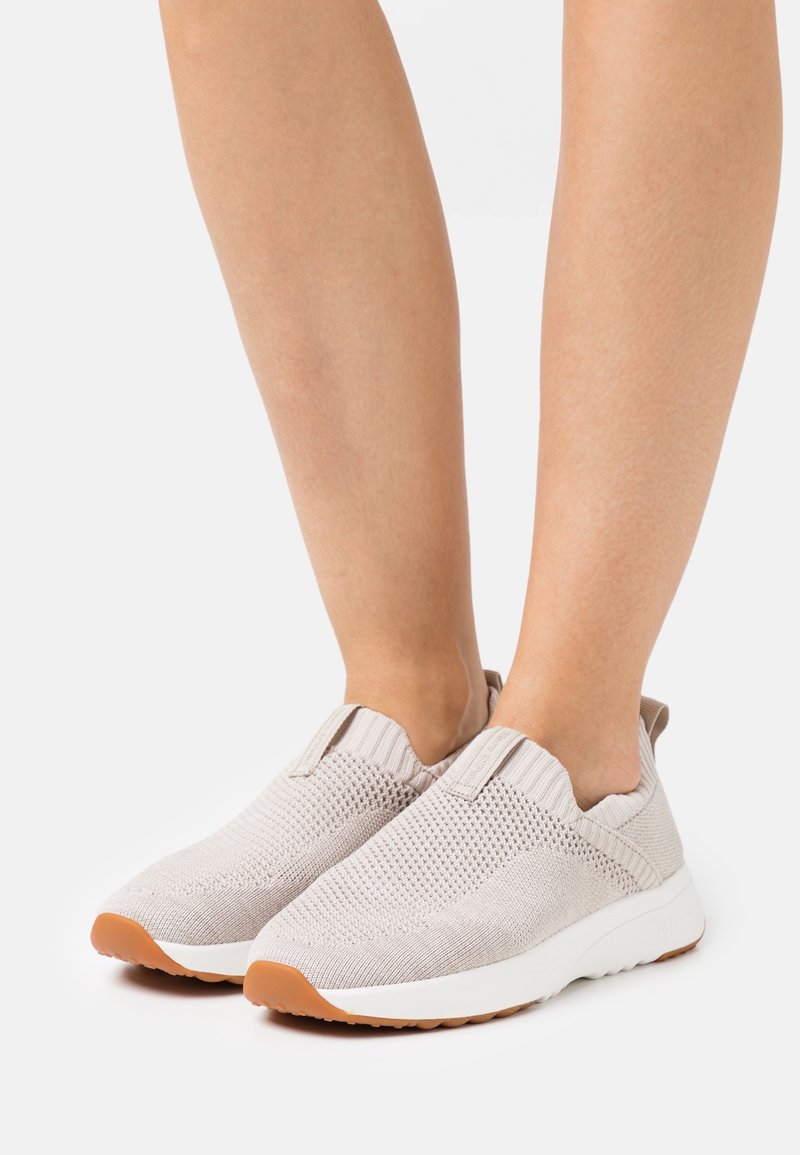 Pieds portant des baskets en maille beige clair à enfiler avec semelles blanches et semelles extérieures en caoutchouc couleur havane, debout sur une surface blanche.