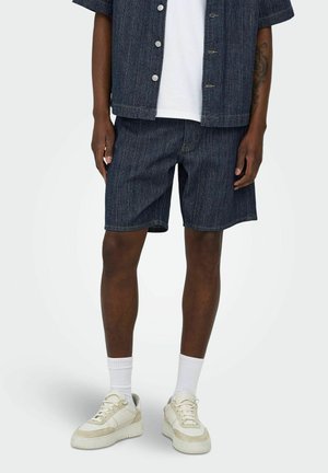 Mann trägt dunkle Denim-Shorts, passendes kurzärmeliges Hemd mit Knöpfen, weiße Socken und beige Sneaker vor neutralem Hintergrund.