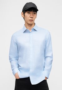 Νέος άνδρας που φοράει ανοιχτό μπλε πουκάμισο με σχέδιο και κουμπιά, μαύρο καπέλο και μαύρο παντελόνι, στέκεται με τα χέρια στις τσέπες μπροστά σε λευκό φόντο.