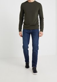 Olivově zelená mikina s dlouhými rukávy kombinovaná s modrými džínami a černými teniskami. Mikina má kulatý výstřih a žebrované manžety.