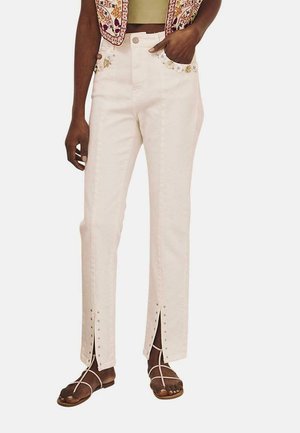 Jeans de tiro alto color beige claro con bolsillos frontales adornados con bordados florales y cortes laterales, con acentos de tachuelas plateadas a lo largo de las costuras.