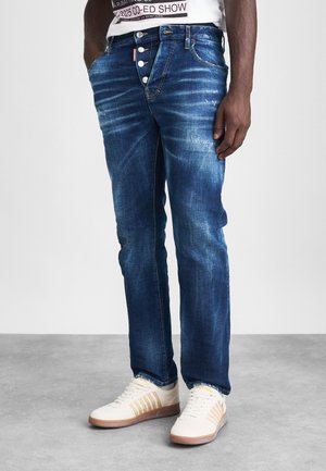 Denim jeans in medium blue with faded details, five-button fly, and classic straight leg. Worn with white and beige sneakers featuring stripes.