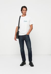Mand iført hvid Calvin Klein t-shirt, mørke jeans, sorte loafers og en sort skuldertaske, stående mod en ensfarvet hvid baggrund.