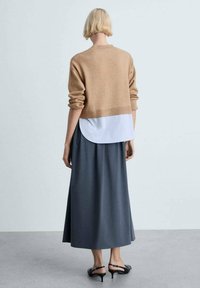 Beiger, kürzer geschnittener Pullover über einer weißen Hemdbluse, kombiniert mit einem dunkelgrauen Maxirock und schwarzen High Heels, der ein geschichtetes Design präsentiert.
