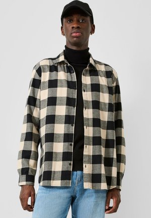 Jonge man met een zwarte pet, een zwarte coltrui, een beige en zwarte geruite blouse met knopen en lichtblauwe spijkerbroek, staand voor een effen achtergrond.