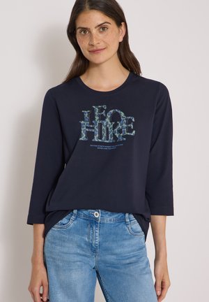 Femme portant un chemisier bleu marine à manches longues avec un texte à motifs "LEO HIKE" et un jean bleu clair sur un fond uni.