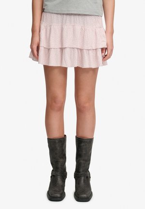 Kvinde iført en lyserød lagdelt mininederdel med subtilt mønster og sorte mellemlange støvler med nittebånd, stående mod en hvid baggrund.