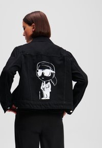 Črna jeans jakna z belim risanim grafičnim motivom na hrbtni strani, ki prikazuje postavo v sončnih očalih in obleki. Klasični ovratnik in gumbi na manšetah.