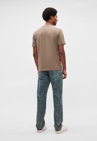 Άνδρας με αφρο μαλλιά που φοράει απλό καφέ κοντομάνικο μπλουζάκι, ξεβαμένα μπλε τζιν και λευκά αθλητικά παπούτσια, όρθιος με την πλάτη γυρισμένη.