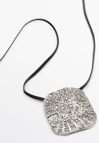 Pendentif en métal argenté avec un design texturé complexe, monté sur un ruban noir. Le pendentif est plat et de forme organique.