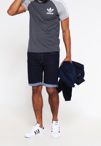 Hombre con una camiseta gris de Adidas, pantalones cortos de mezclilla oscuros y doblados, y zapatillas blancas, sosteniendo una chaqueta azul marino doblada en su mano derecha.