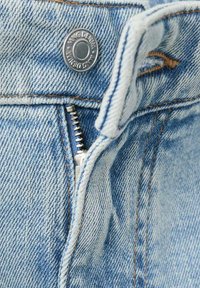 Denim azul claro con un botón de tono cobre, una cremallera metálica y costuras visibles. Primer plano del detalle de la cintura.