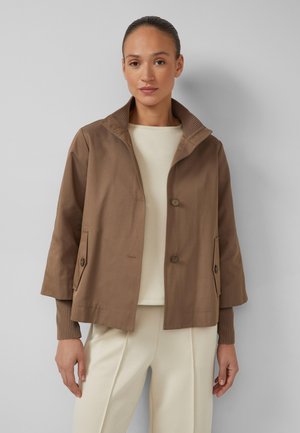 Woman wearing a brown cropped jacket with buttoned pockets over a cream top and cream pants, standing against a plain light gray background.