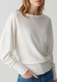 Weißer, leichter Strickpullover mit weitem Ausschnitt, langen Dolmanärmeln und geripptem Saum, kombiniert mit blauen Jeans. Weiche Textur und lässige Passform.