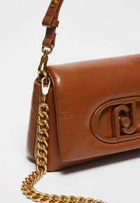 Sac à main en simili cuir marron avec une bandoulière en chaînes dorées et un grand logo brillant sur le devant. Texture lisse avec une forme structurée.