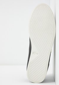 Semelle en caoutchouc blanche d'une basket noire avec des lignes de bande de roulement diagonales et un petit logo de crocodile près de la voûte plantaire.