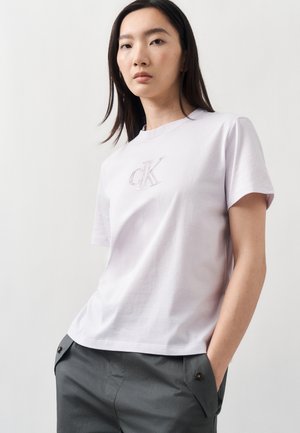 Joven mujer con cabello liso y oscuro, llevando una camiseta blanca de Calvin Klein y pantalones grises, con las manos en los bolsillos, fondo neutro.