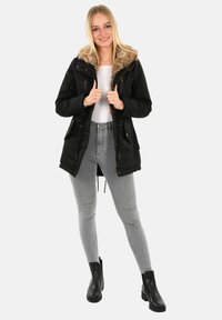 Junge Frau in einer schwarzen, mit Fell gefütterten Parka, grauen Skinny Jeans und schwarzen Stiefeletten, die mit den Daumen in die Jacke eingehakt steht.