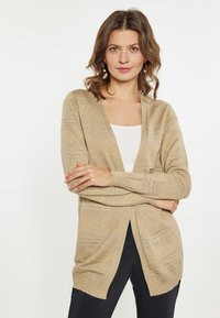 Cardigan beige realizzato in tessuto morbido e testurizzato, con scollo a V, apertura frontale e dettagli a righe sottili, abbinato a una maglietta bianca e pantaloni scuri.
