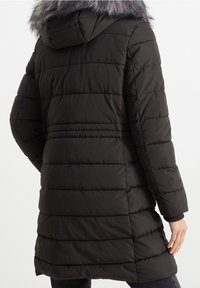 Svart pufferjacka med ett horisontellt kviltat mönster och en avtagbar grå pälshuva. Långa ärmar och en design som går till knäna.