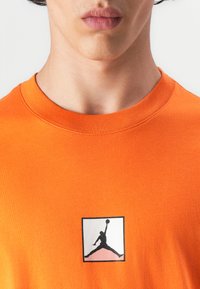Orange bomulds t-shirt med et centreret sort-hvidt Air Jordan-logo i en firkant. Glat tekstur med rund hals.