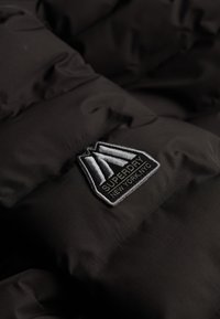 Sort pufferjakke stof med quiltet design. Indeholder en fremtrædende broderet Superdry-logo mærkat i grå og hvid.