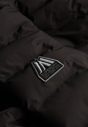 Zwarte pufferjasstof met doorgestikt ontwerp. Voorzien van een opvallend geborduurd Superdry-logo label in grijs en wit.