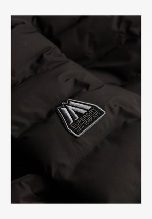 Zwarte pufferjasstof met doorgestikt ontwerp. Voorzien van een opvallend geborduurd Superdry-logo label in grijs en wit.