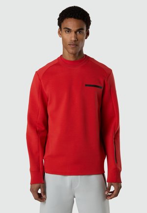 Homme portant un sweat-shirt à manches longues rouge avec une fermeture éclair noire sur la manche et un détail noir sur la poitrine, associé à un pantalon gris clair.